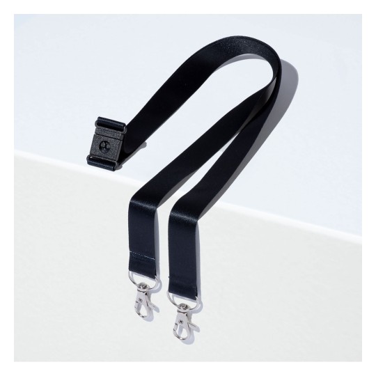 Kuvassa on musta hihna, jonka toisessa päässä on yksi klipsi ja toisessa päässä kaksi metalliklipsiä, lepäämässä vaalealla pinnalla.