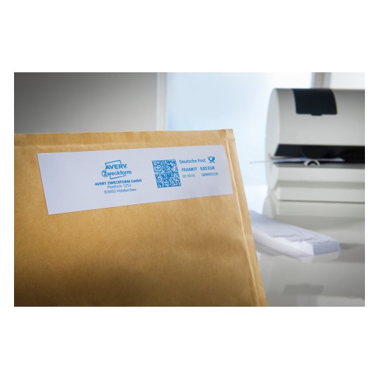 The image shows a brown envelope with a white label featuring company details, a QR code, and postage information. In the background, a printer and other office materials are visible.