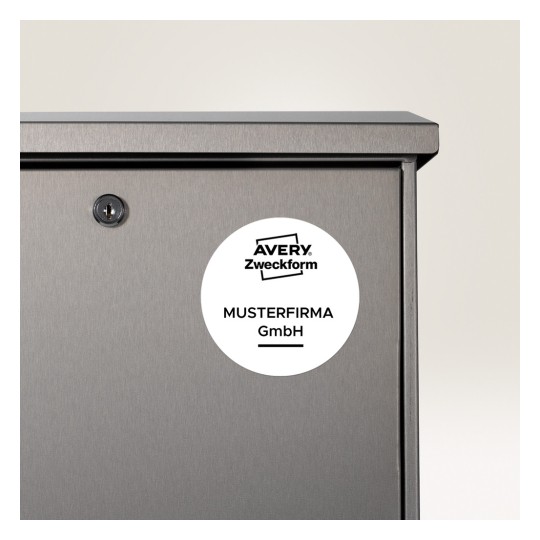 Kuvassa näkyy harmaan metalliesineen lähikuva, jossa on avaimenreikä ja pyöreä etiketti. Etiketissä on teksti 'AVERY Zweckform MUSTERFIRMA GmbH', mikä viittaa siihen, että kyseessä voi olla säilytysyksikkö tai lokero.