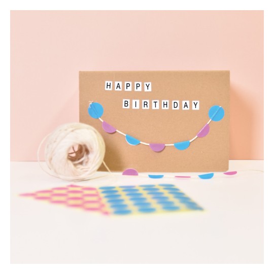 Kuva esittää ruskean kortin, jossa lukee 'HAPPY BIRTHDAY'. Kortin poikki roikkuu koristeellinen sinisistä ja violetista ympyröistä tehty viiri. Etualalla on rulla narua sekä kuviollisia papereita pinkissä, keltaisessa ja sinisessä värissä.