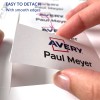 Kuvassa näkyy käsi irrottamassa tarraa arkista, jossa on nimi 'Paul Meyer' ja 'Avery' -logo, ja muistiinpano, joka osoittaa, että se on helppo irrottaa sileiden reunojen ansiosta.