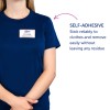 Kuvassa näkyy henkilö, joka on pukeutunut siniseen paitaan, jossa on nimilappu. Lappu lukee 'B. Carlos, Markkinointijohtaja'. Teksti osoittaa, että lappu on itsekiinnittyvä, ja se voi tarttua vaatteisiin sekä irrota helposti jättämättä jäämiä.