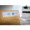 The image shows a brown envelope with a white label featuring company details, a QR code, and postage information. In the background, a printer and other office materials are visible.